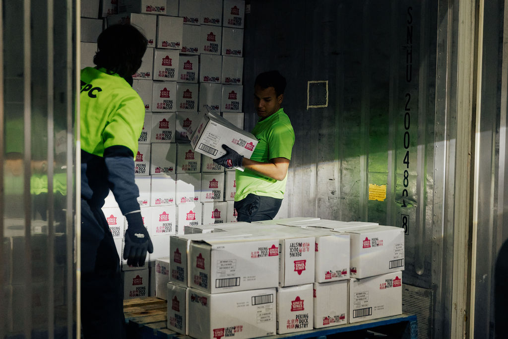Cold storage warehouse with stacked wooden crates of fresh apples and other produce beneath ceiling-mounted refrigeration units.