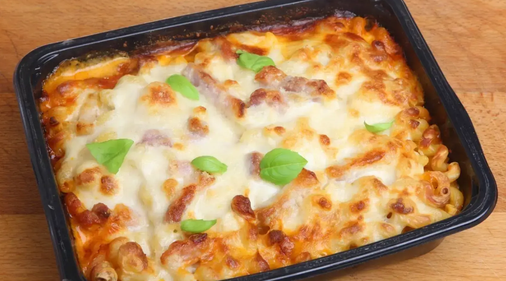 Baked pasta meal in a black tray, topped with melted golden cheese and fresh basil leaves.